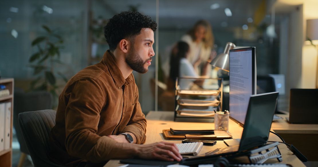 Un homme travaille seul à un bureau la nuit, concentré sur son ordinateur, avec des lumières de bureau derrière.