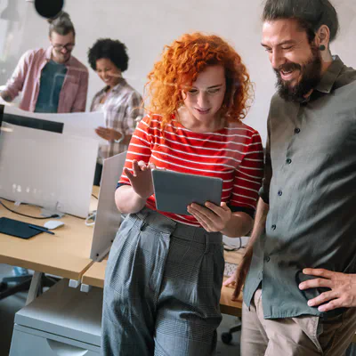 Deux personnes debout regardent une tablette dans un bureau, tandis que des collègues travaillent derrière.