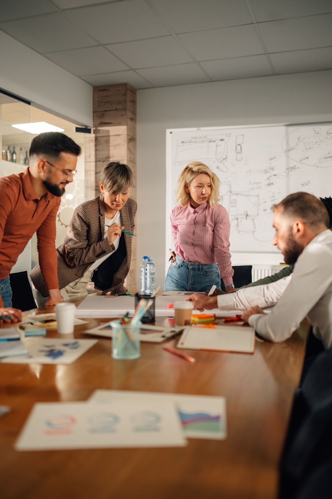 Quatre collègues réunis autour d’une table avec des documents, discutant devant un tableau blanc couvert de schémas.