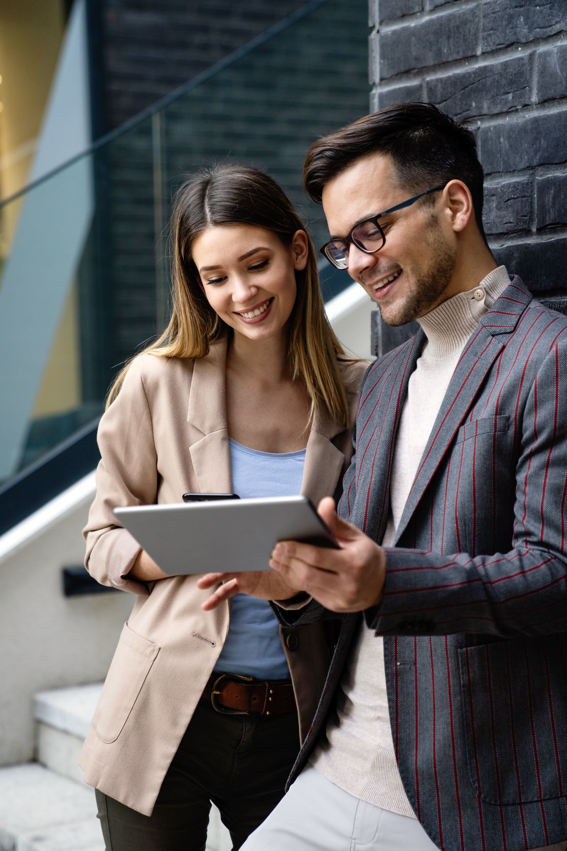 Deux collègues sourient en regardant ensemble une tablette, debout près d’un escalier moderne de bureau et d’un mur.